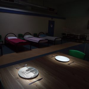 neatly made beds in the darkened room at the Julian Trust Night Shelter, in the foreground a table is laid for a meal onto one of the empty plates the image of feet walking along a street is projected