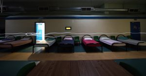 a photograph from Shelter a poetry and film project working with Julian Night Trust. It shows a darkened room with five beds neatly made and pushed against the wall with an open bathroom door out of which a bright blue light emanates